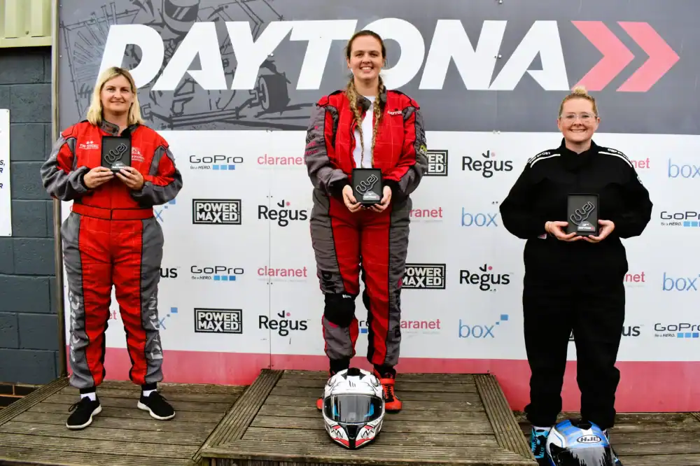 Three women in racing suits stand on a podium holding trophies in front of a Daytona backdrop, indicating a motorsport event. Helmets rest on the ground in front of them.