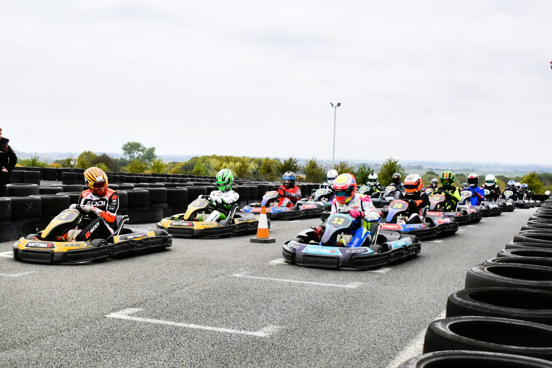 A group of go-kart drivers wearing helmets start a race on an outdoor track lined with black tyres.