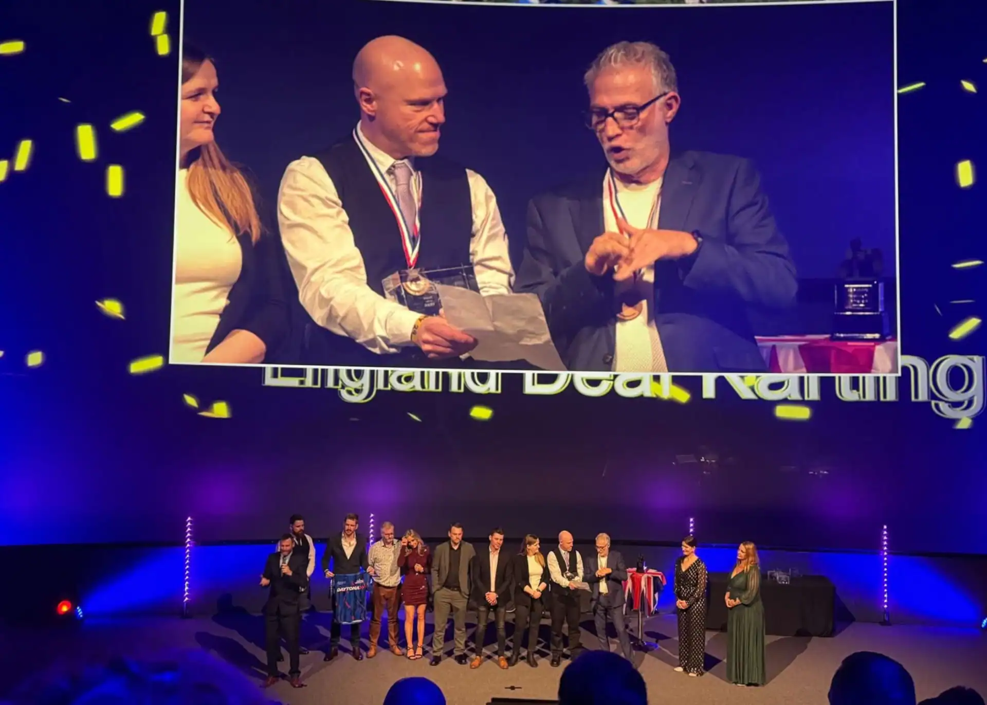 A group of people stand on a stage at an awards event, with two men shown on a large screen above, one holding a medal and paper.