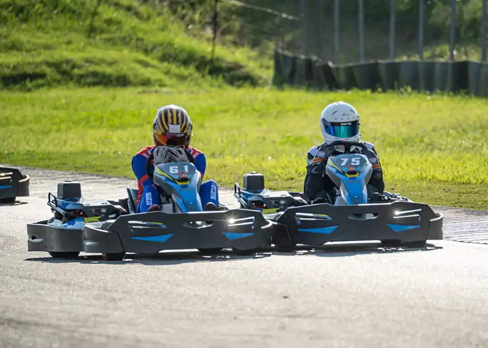 Two go-kart drivers, numbered 61 and 75, race side by side on a tarmacked outdoor track with grassy surroundings.