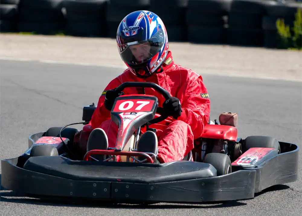 Person wearing a red racing suit and helmet driving a go-kart numbered 07 on an outdoor circuit with tyres in the background.