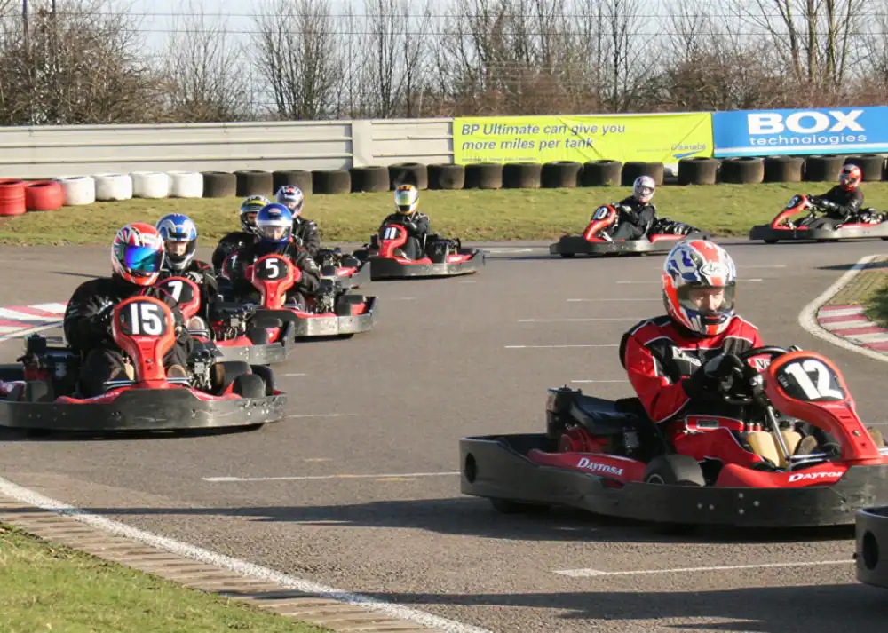 Eight people drive go-karts round a bend on an outdoor track, all wearing helmets and racing suits, with tyre barriers and trees in the background.