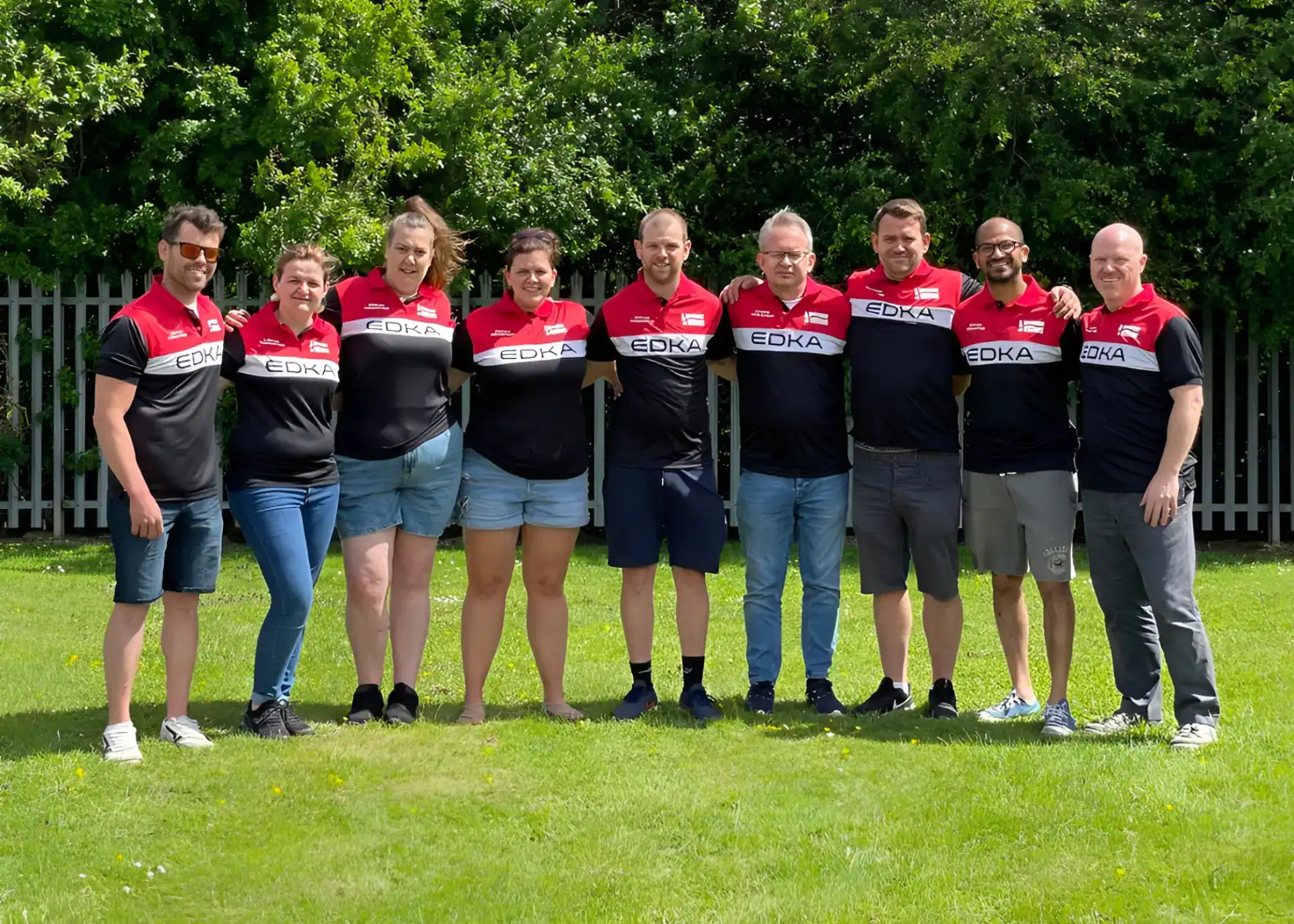 A group of nine people in matching black, red, and white shirts stand together on grass with trees and a metal fence in the background.