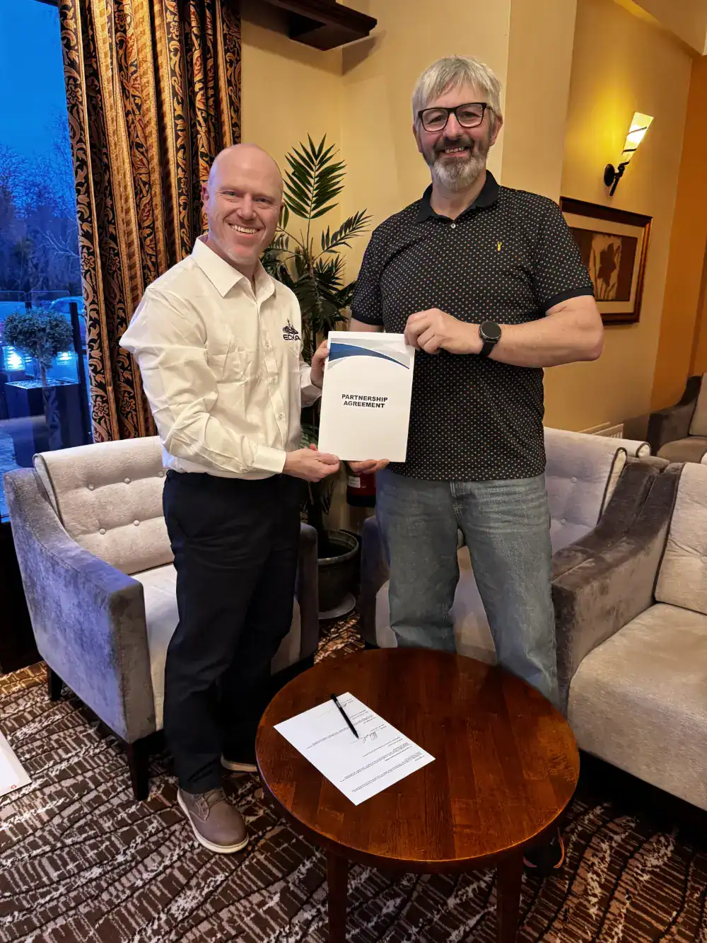 Two men stand indoors next to a table, holding a document titled "Partnership Agreement," with a signed paper and pen on the table in front of them.