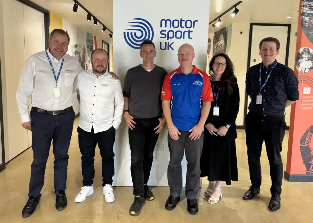 Six people stand in a row, smiling, in front of a Motor Sport UK sign in an indoor setting.