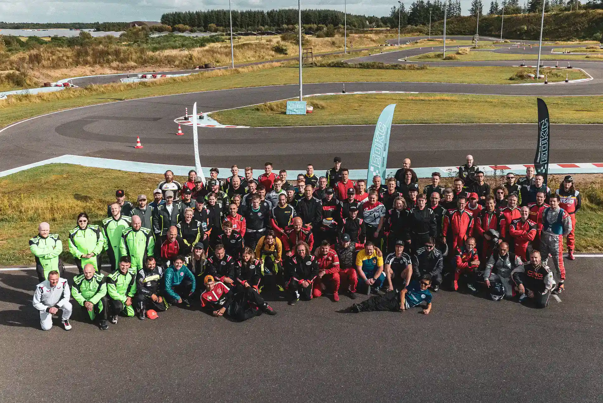 A large group of people in racing suits pose together on an outdoor go-kart track with green grass and winding tarmac in the background.