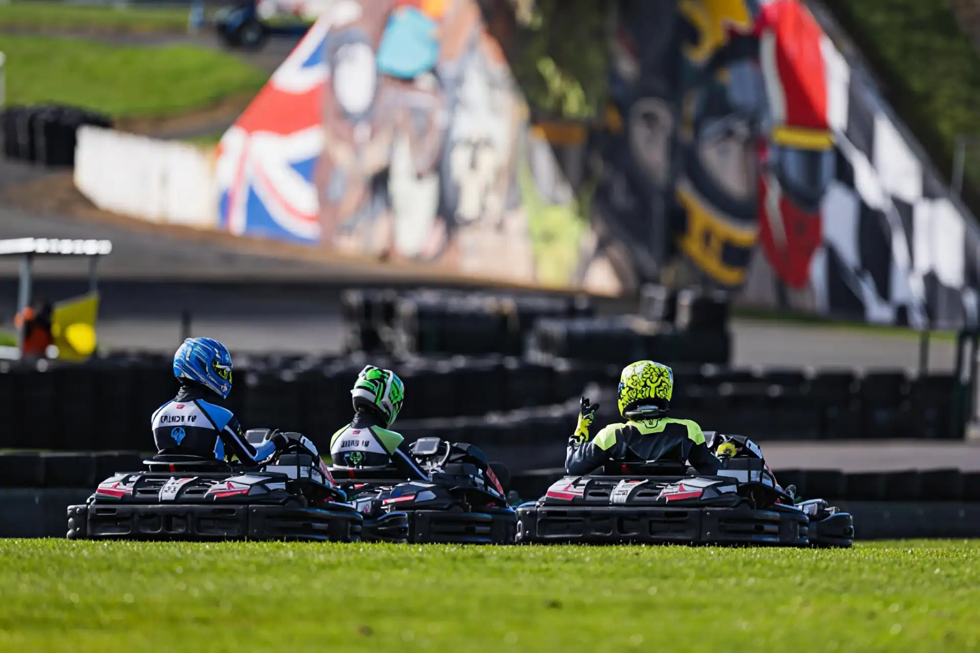 Three go-kart drivers in racing gear drive on a track with grass in the foreground and a colourful mural on the wall in the background.