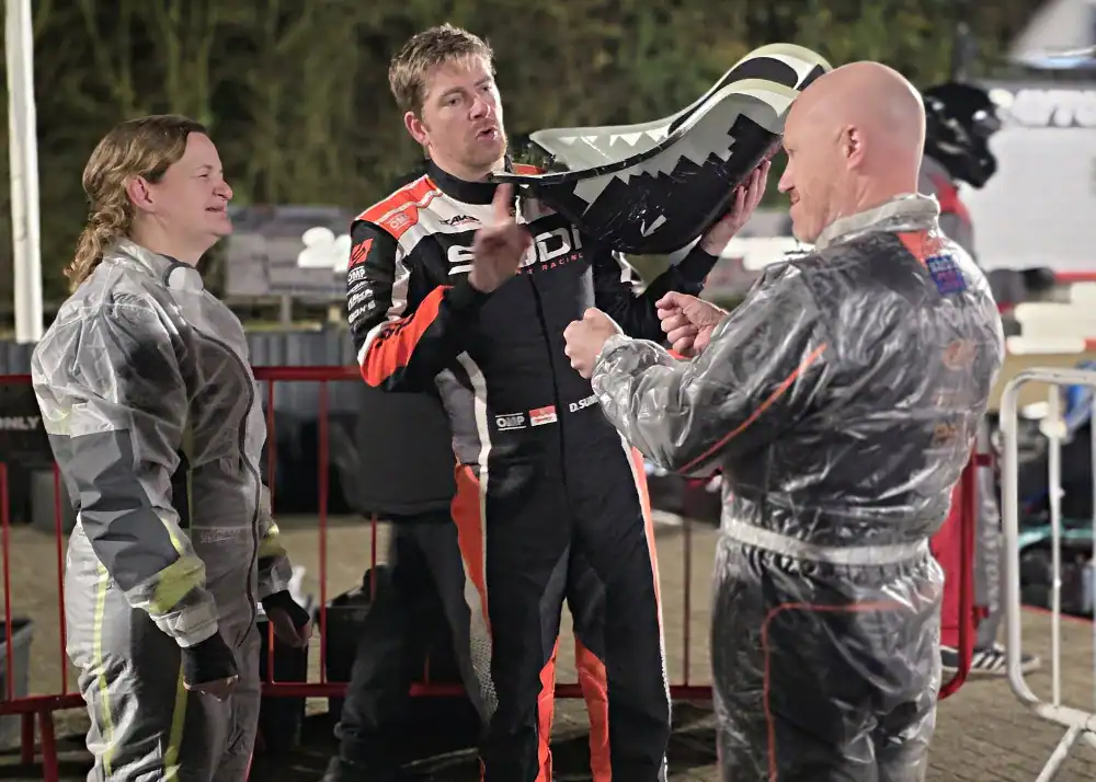 Three people in racing suits stand together; one holds a go-kart seat whilst talking to the others. A barrier and blurred background are visible.