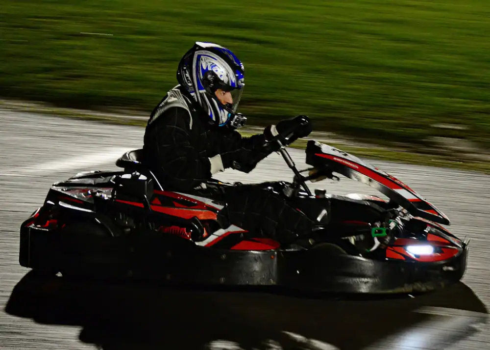 Person wearing a helmet and racing suit driving a go-kart on an outdoor track at night, with blurred background indicating motion.