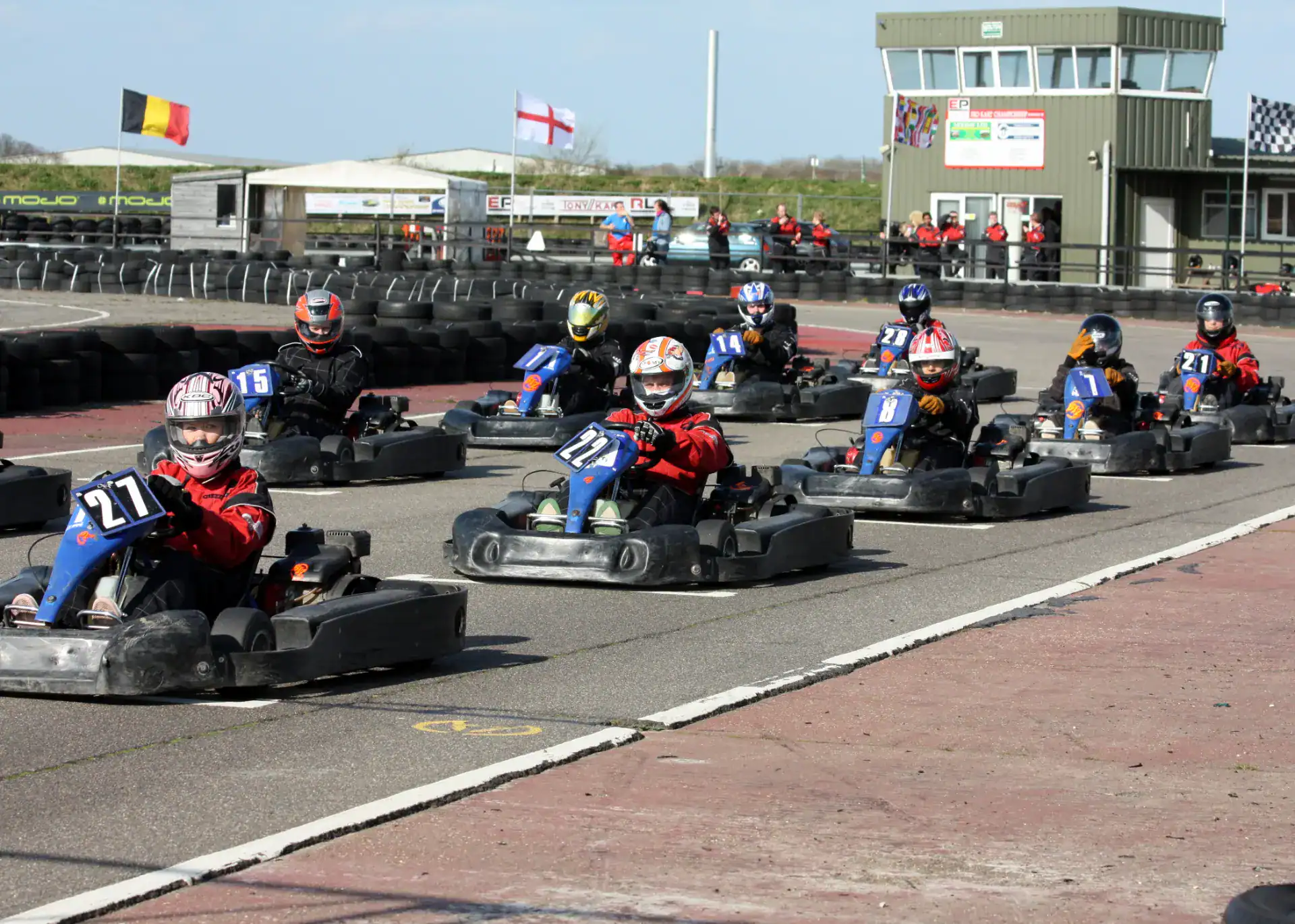 A group of people wearing helmets drive go-karts on a racetrack, with a control tower and flags visible in the background.