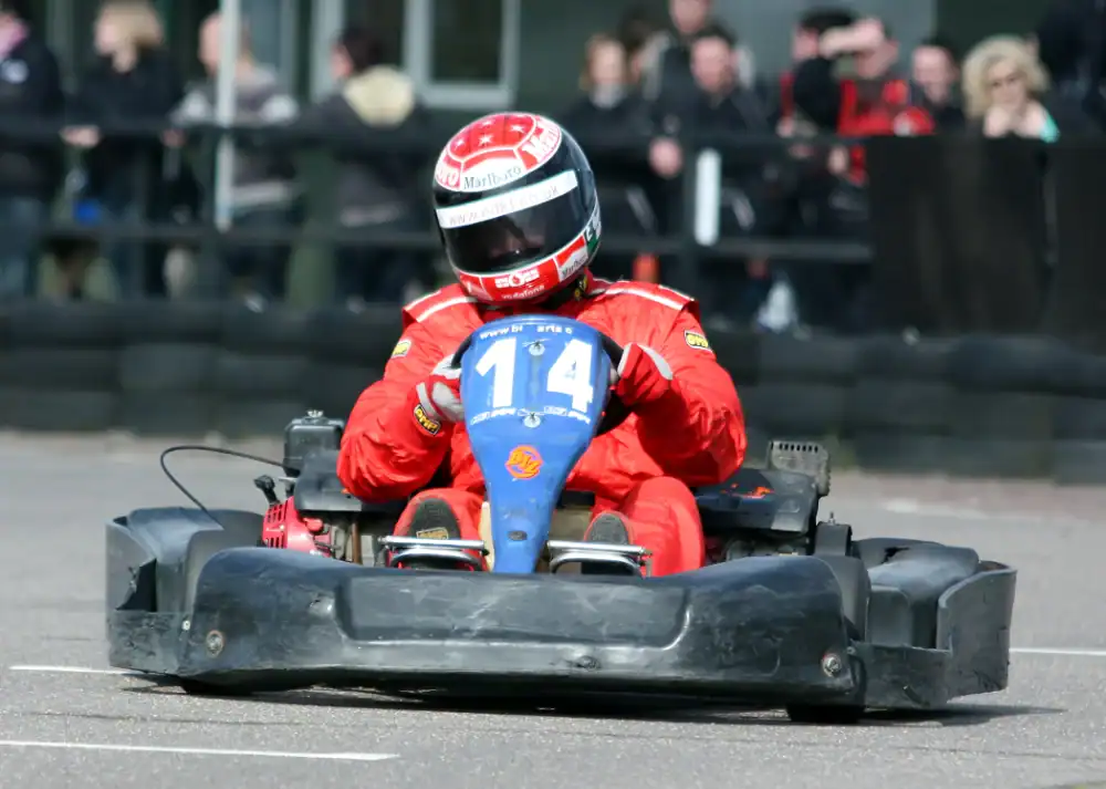 A person in a red racing suit and helmet drives a go-kart with the number 114 on a tarmacked track, with spectators in the background.