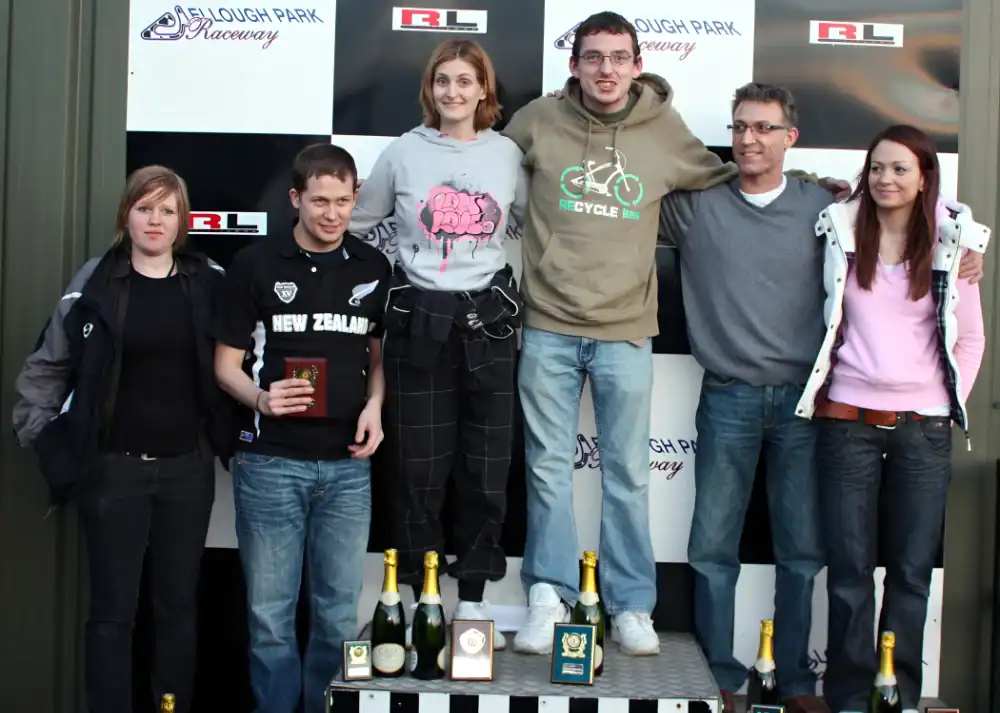 Six people stand on and around a podium at an indoor karting event, holding plaques and bottles, with a chequered backdrop behind them.