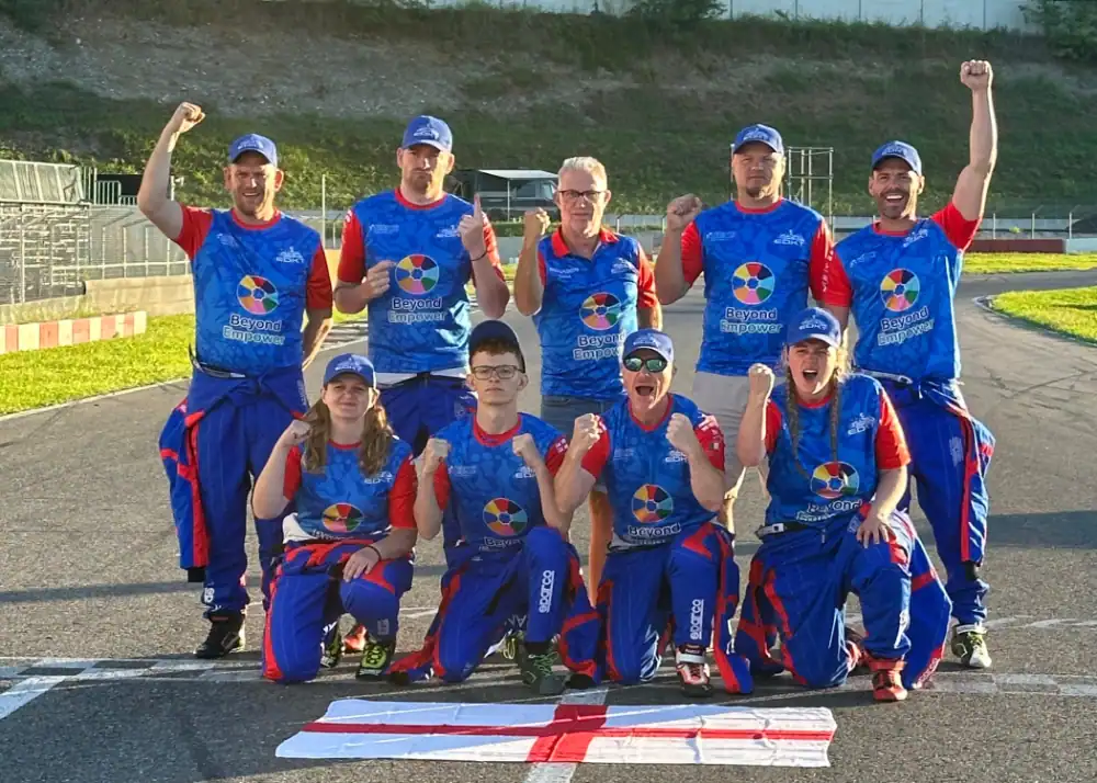 Nine people in matching blue and red uniforms pose cheering on a race track, standing and kneeling behind an England flag laid flat on the ground.