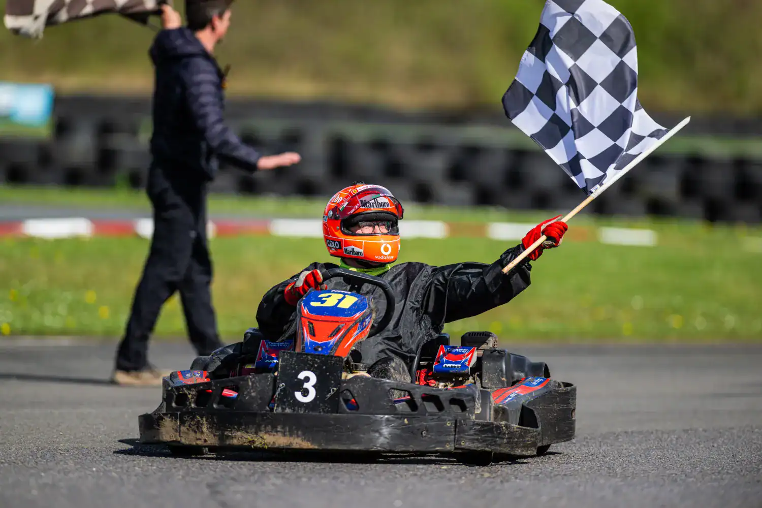 Go-kart driver wearing a red helmet holds a chequered flag whilst crossing the finishing line on an outdoor track, with a person in the background holding another flag.