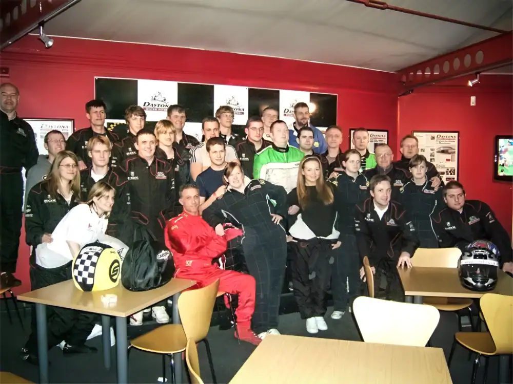 A group of people in racing suits pose together indoors next to tables and helmets, with race-related posters on the red wall behind them.
