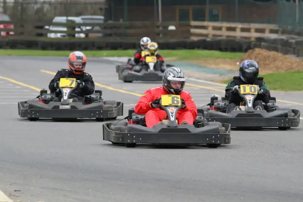 Four people in helmets and racing suits drive go-karts on an outdoor track, with visible numbers 6, 7, and 10 on the karts.