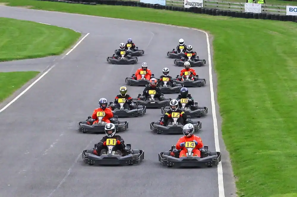 A group of go-kart drivers wearing helmets and numbered vests race on an outdoor track, closely following each other round a bend.