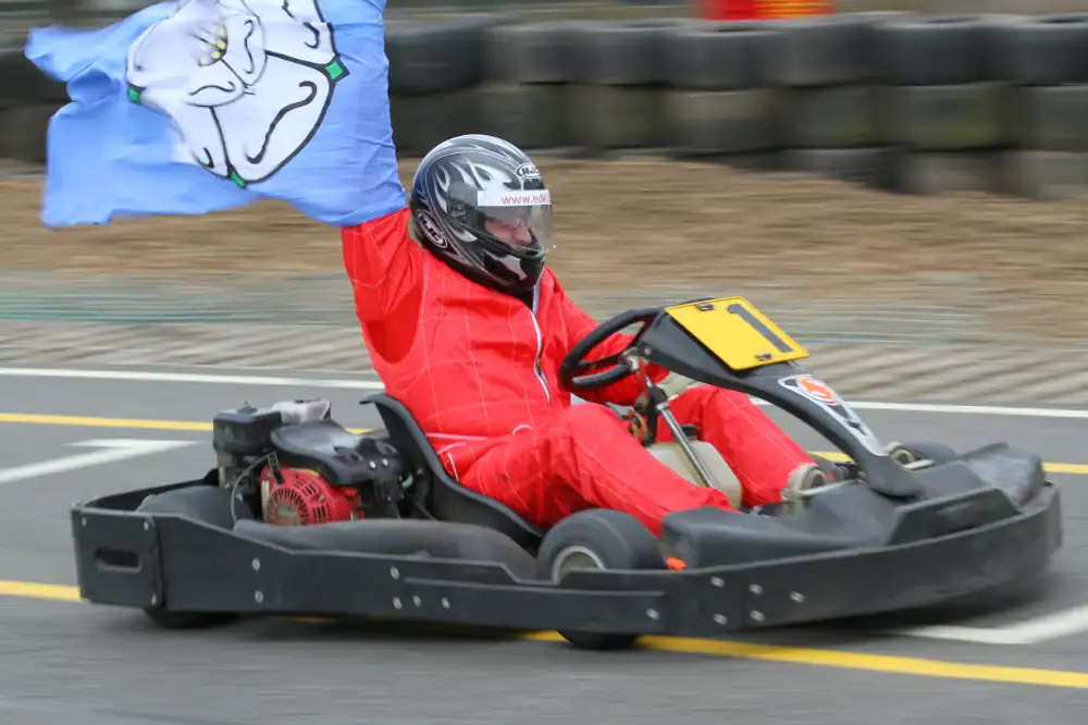 Person in a red racing suit and helmet driving a go-kart whilst holding up a blue flag with a cartoon face on it, on an outdoor track.