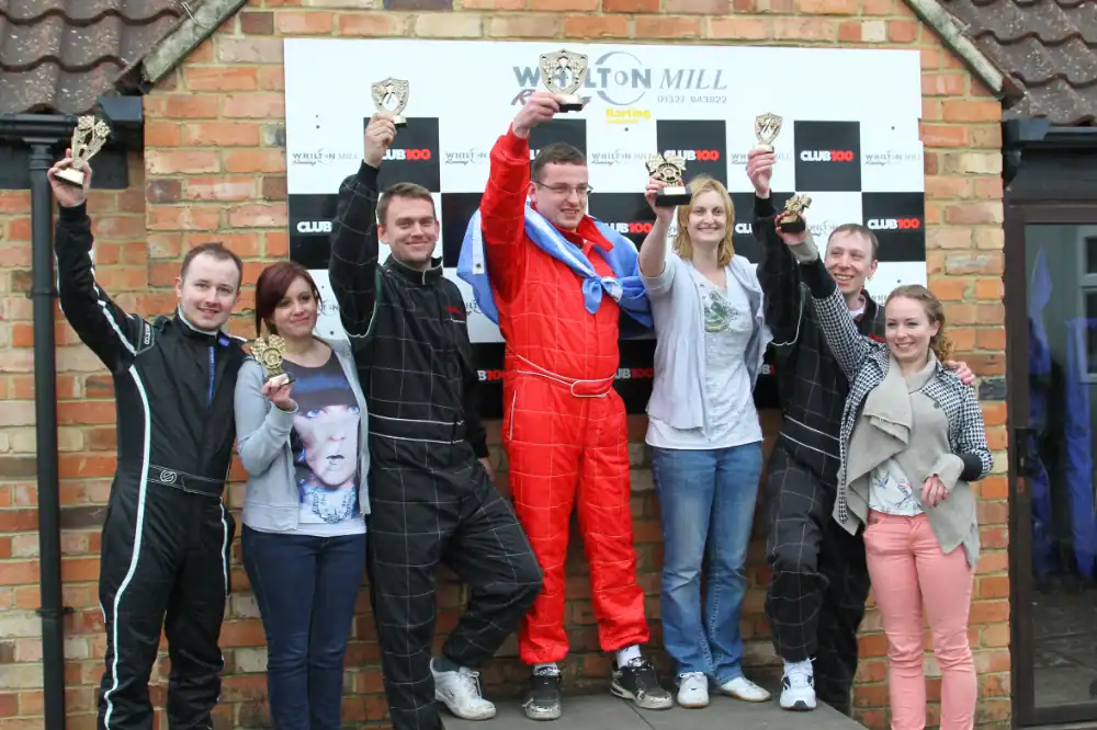 Eight people stand in front of a brick wall and racing backdrop, some in racing suits, holding up trophies and posing for a group photo.