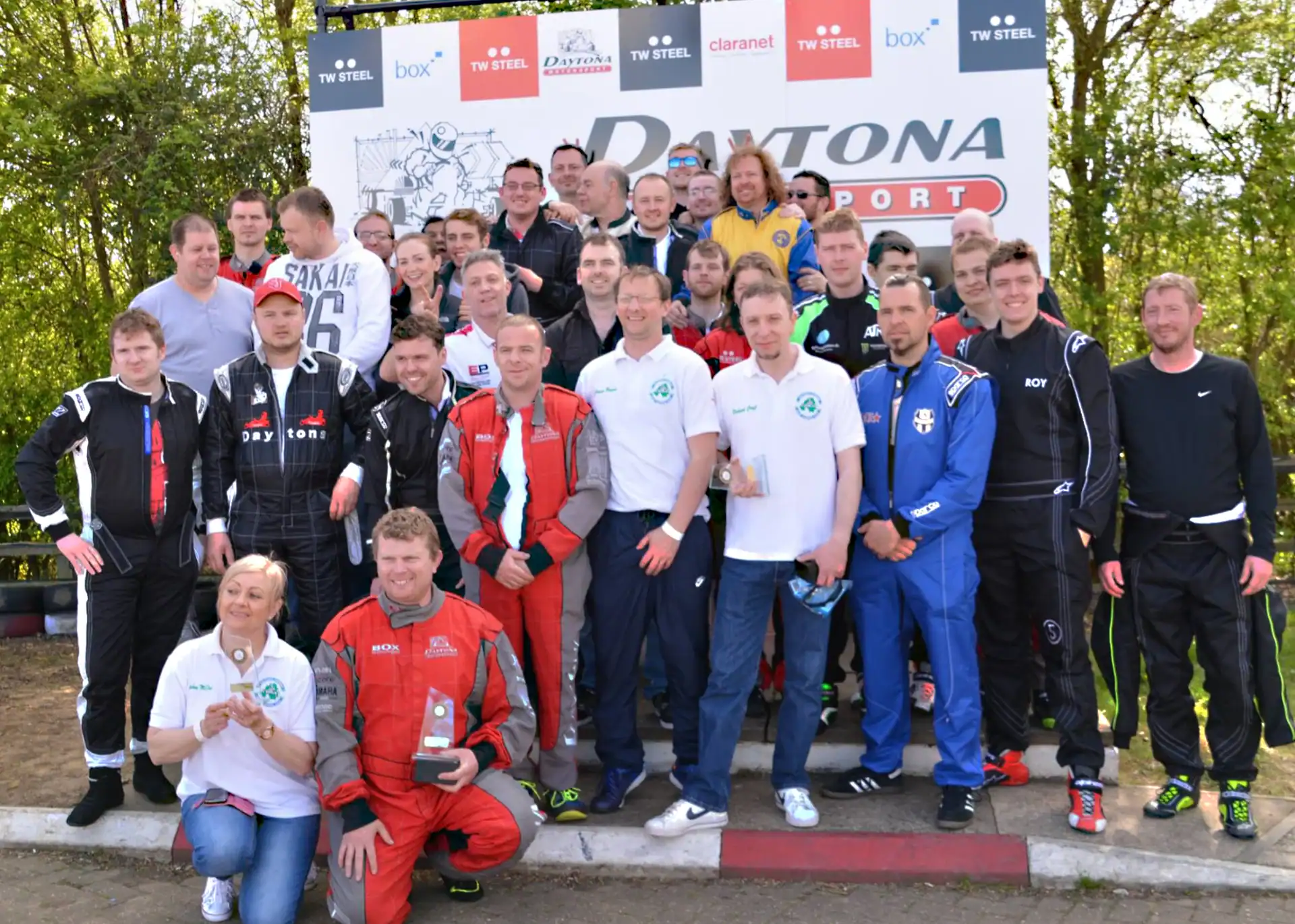 A group of people, some in racing suits and others in casual clothing, posing together in front of a Daytona Motorsport sign outdoors.