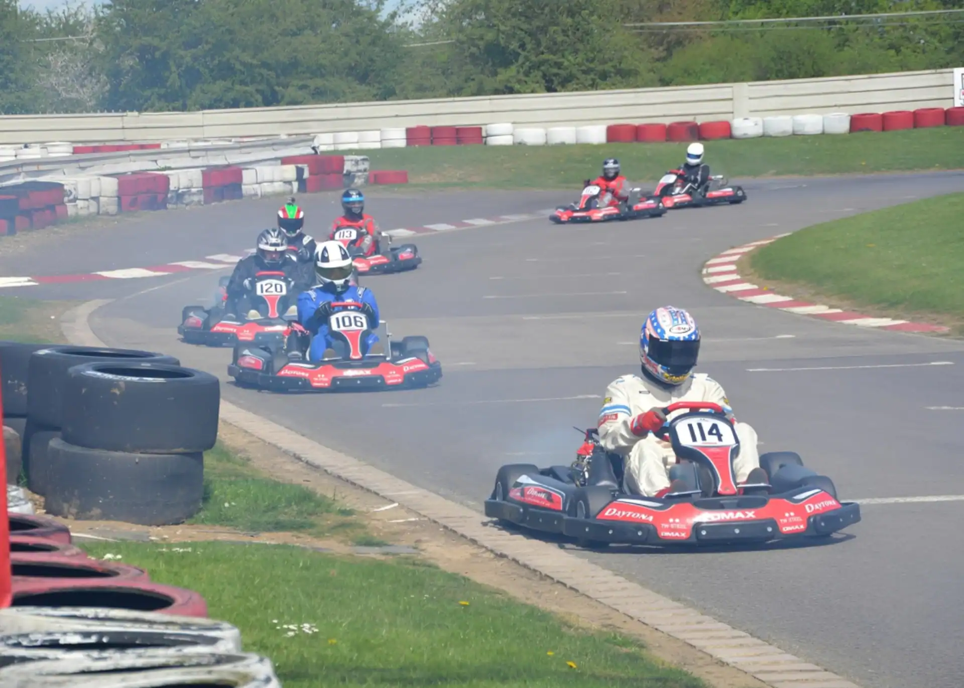 Several go-karts, each with visible numbers, race around a curved outdoor track bordered by red and white barriers on a sunny day.