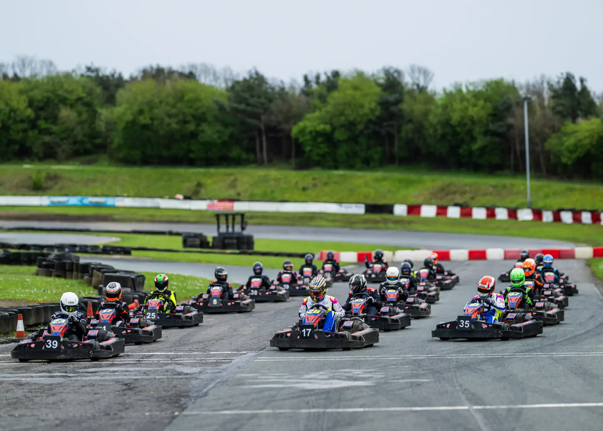 A group of go-kart drivers wearing helmets race on an outdoor track surrounded by grass, trees, and red-and-white barriers.