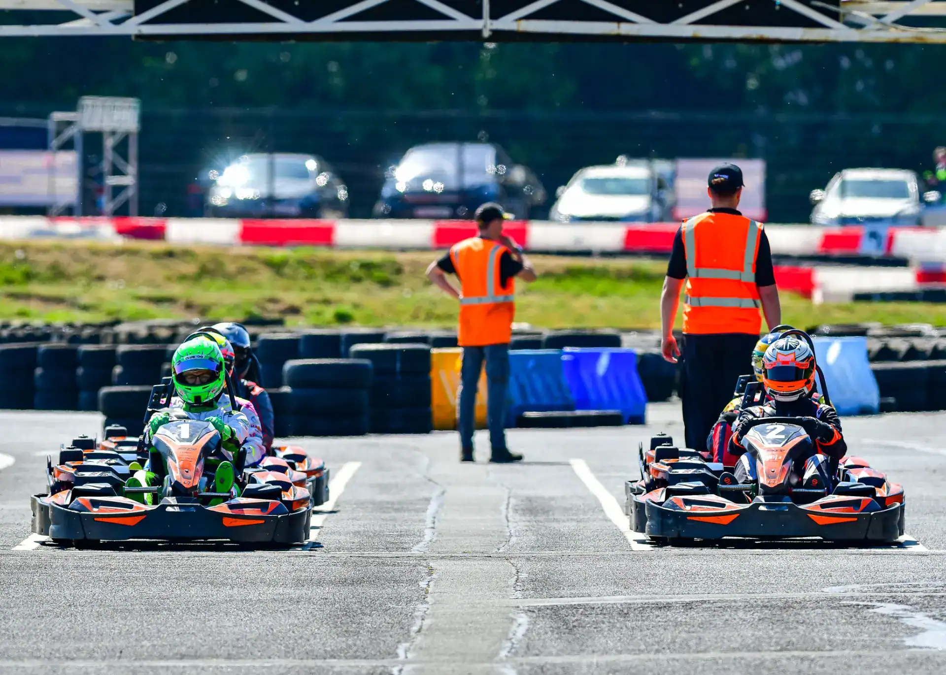 Two go-kart drivers wearing helmets race on a track, watched by two people in orange safety vests. Parked cars and tyre barriers are visible in the background.