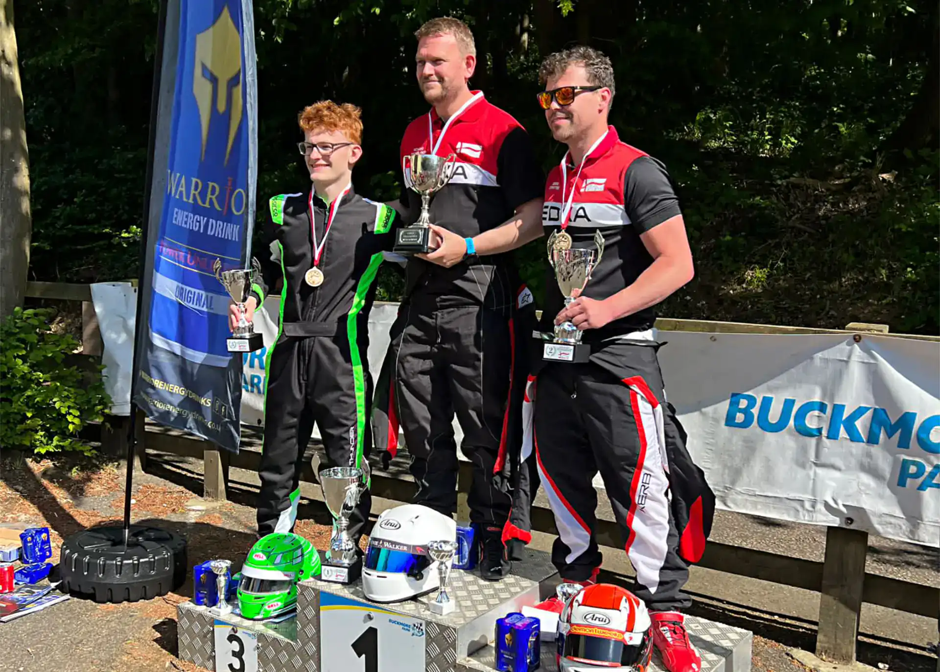 Three men in racing suits stand on a winners’ podium holding trophies and medals, with racing helmets and drinks in front of them at an outdoor karting event.