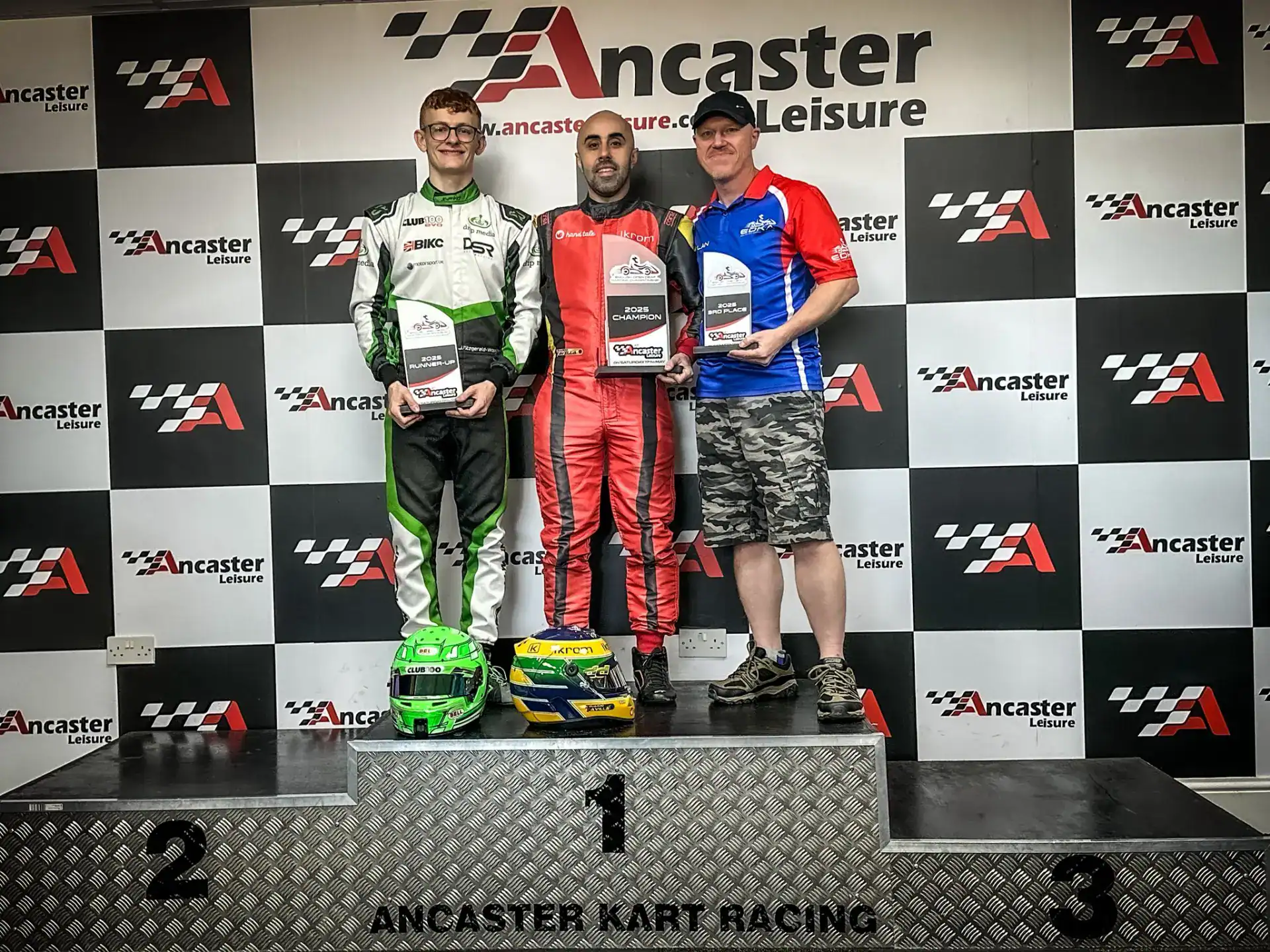 Three men stand on a podium holding trophies at Ancaster Kart Racing, each on a different step marked 1, 2, and 3, with a branded backdrop behind them.