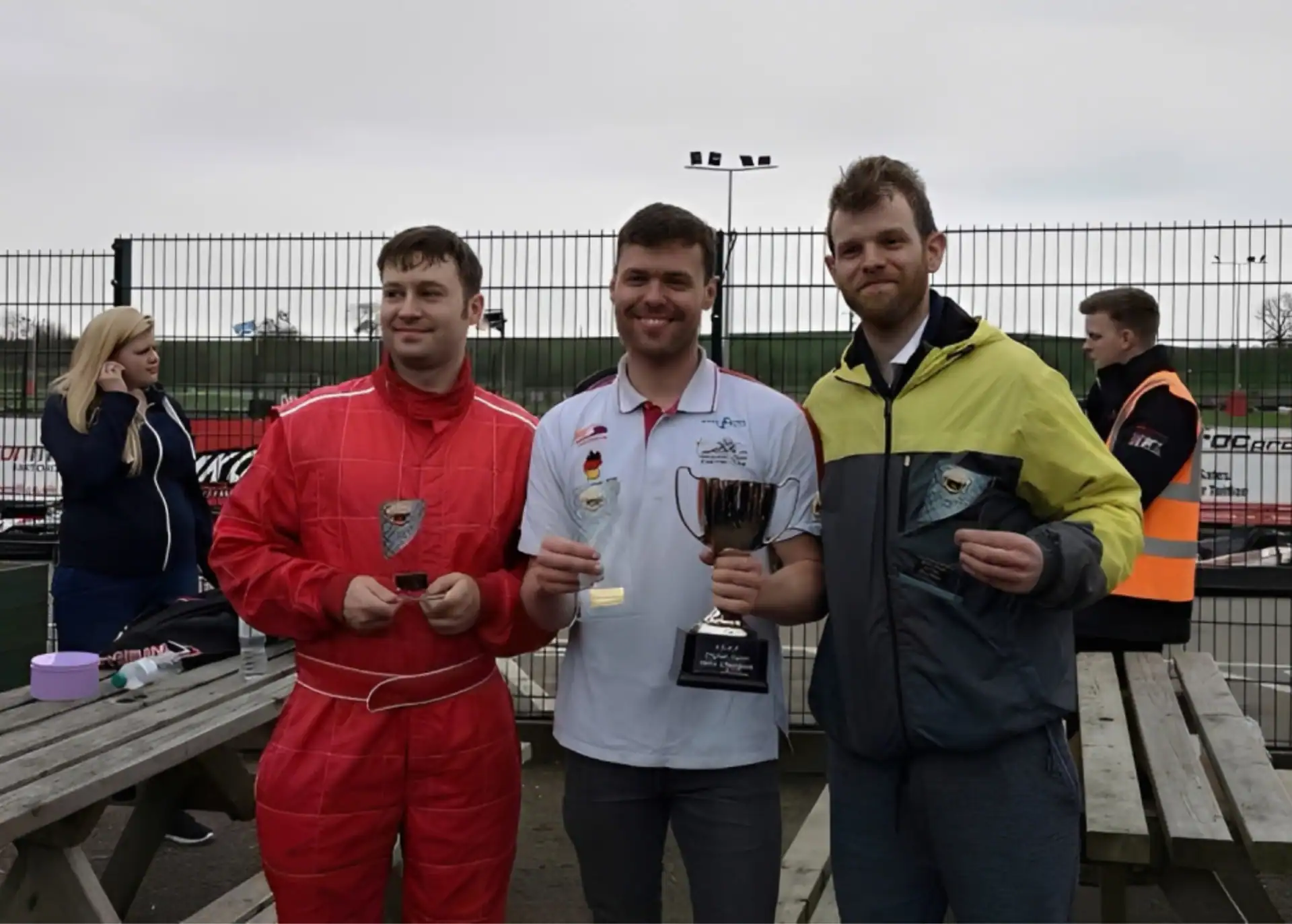 Three men pose outside by a fence; one holds a trophy, another wears a red racing suit, and the third holds a plaque. Go-karts and people are visible in the background.
