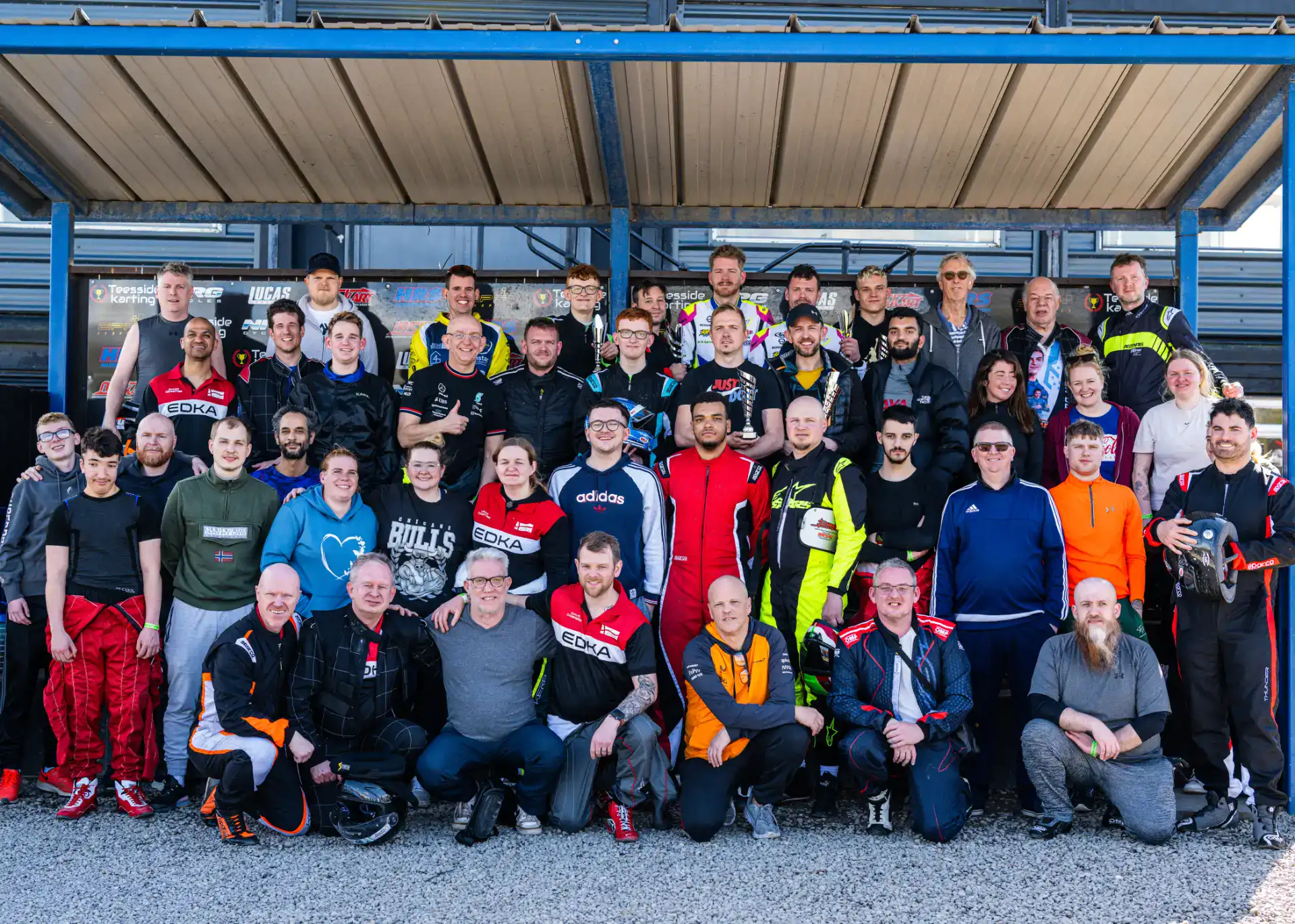 A large group of people in racing suits and casual clothing pose together for a group photo outside a building with a metal roof.