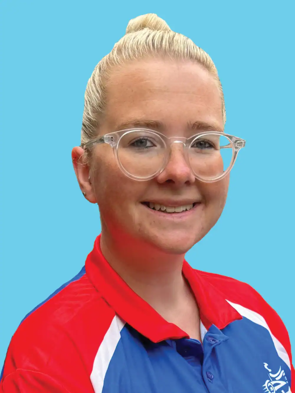 A person with blonde hair in a bun, wearing clear glasses and a red, blue, and white collared shirt, smiles in front of a plain blue background.