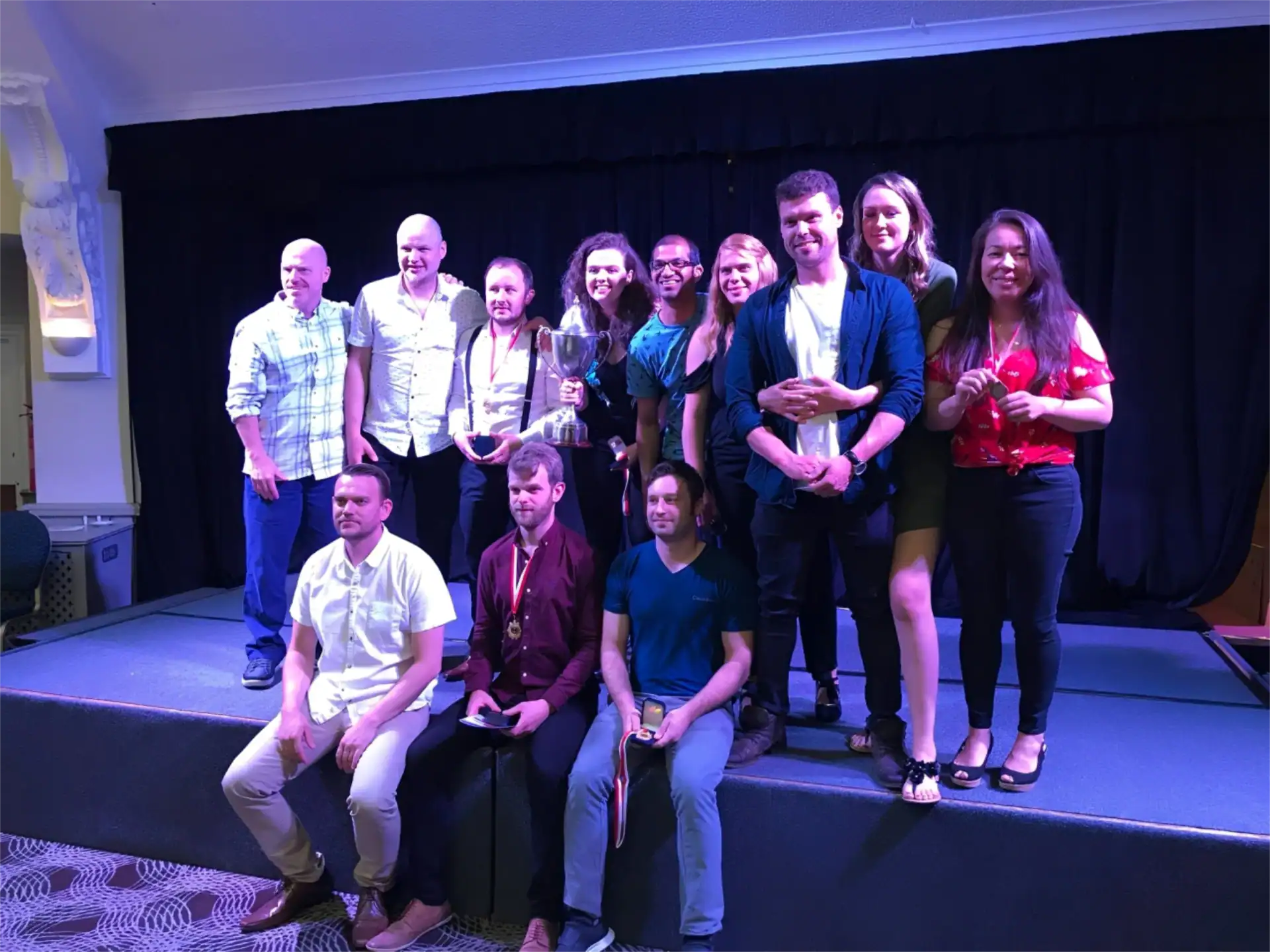 A group of thirteen people, some holding a trophy, pose together on a stage with a black curtain backdrop.