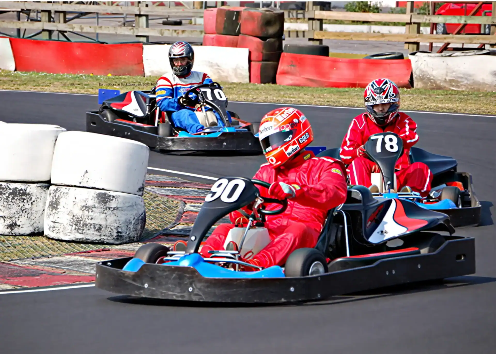 Three people in racing suits and helmets drive go-karts numbered 90, 78, and 70 on an outdoor track with tyre barriers.