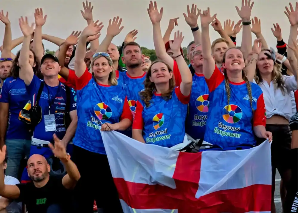 A group of people in blue and red shirts stand together holding a flag, looking and reaching upwards with raised hands, some smiling.