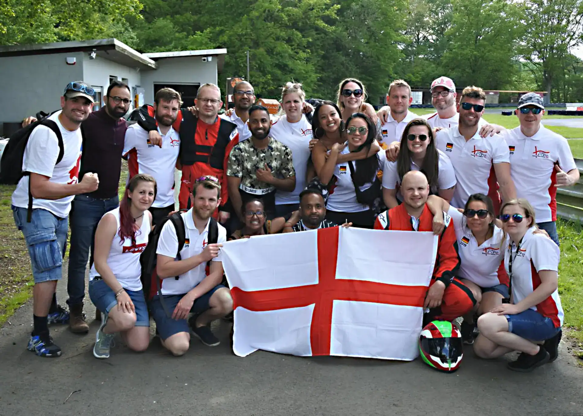 A group of people pose outdoors, some wearing matching white shirts, with a large England flag displayed at the front and trees in the background.