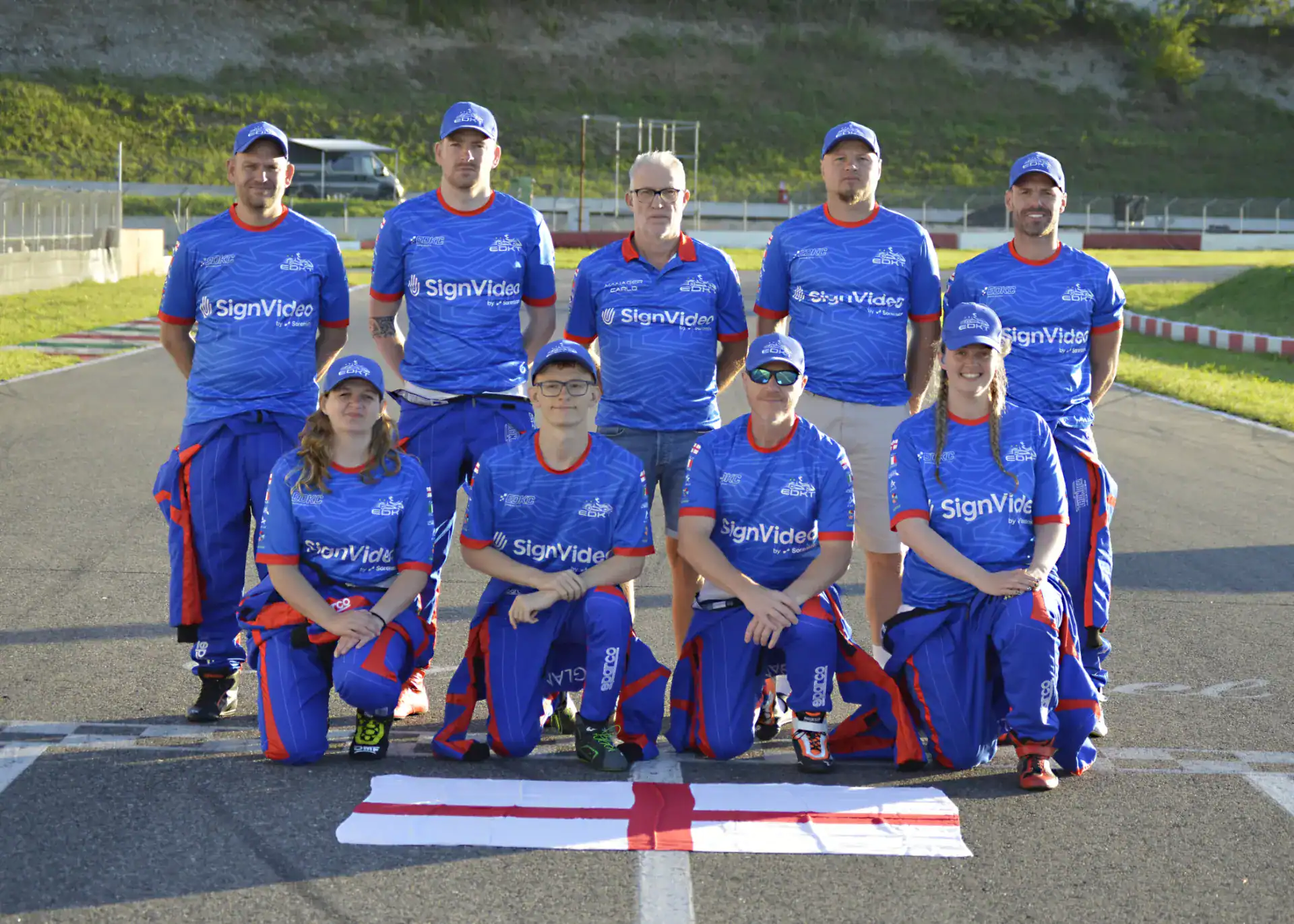 Nine people in blue sports kits pose for a team photo on an outdoor track, with a red and white flag laid on the ground in front of them.