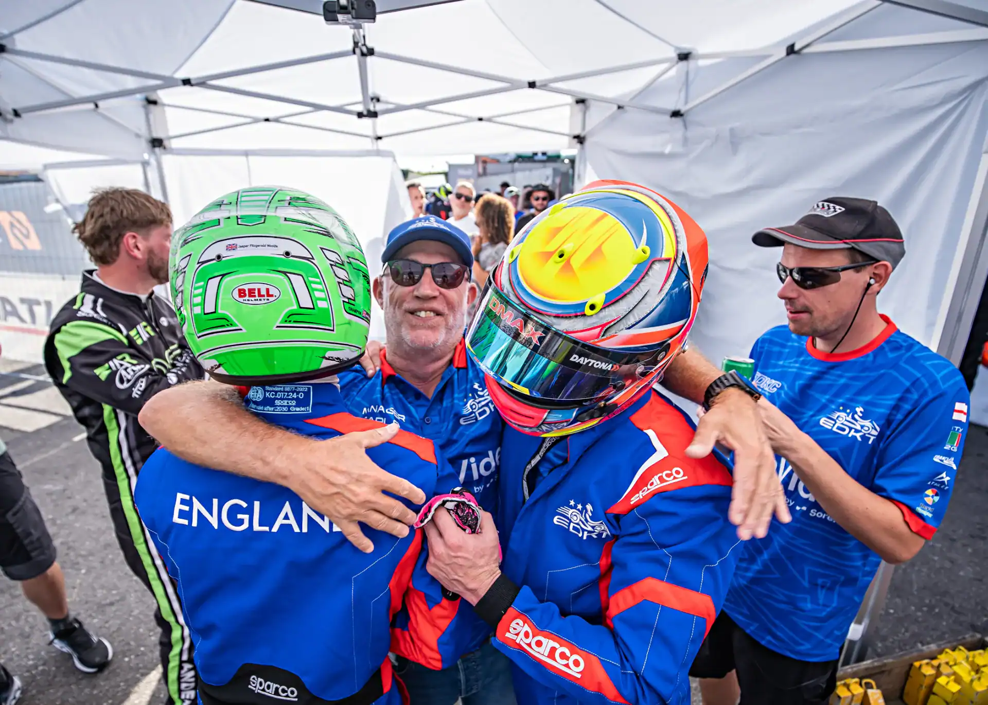 Two racing car drivers in blue suits and helmets embrace whilst a man in sunglasses and a cap smiles and joins the hug under a canopy, with another man standing nearby.