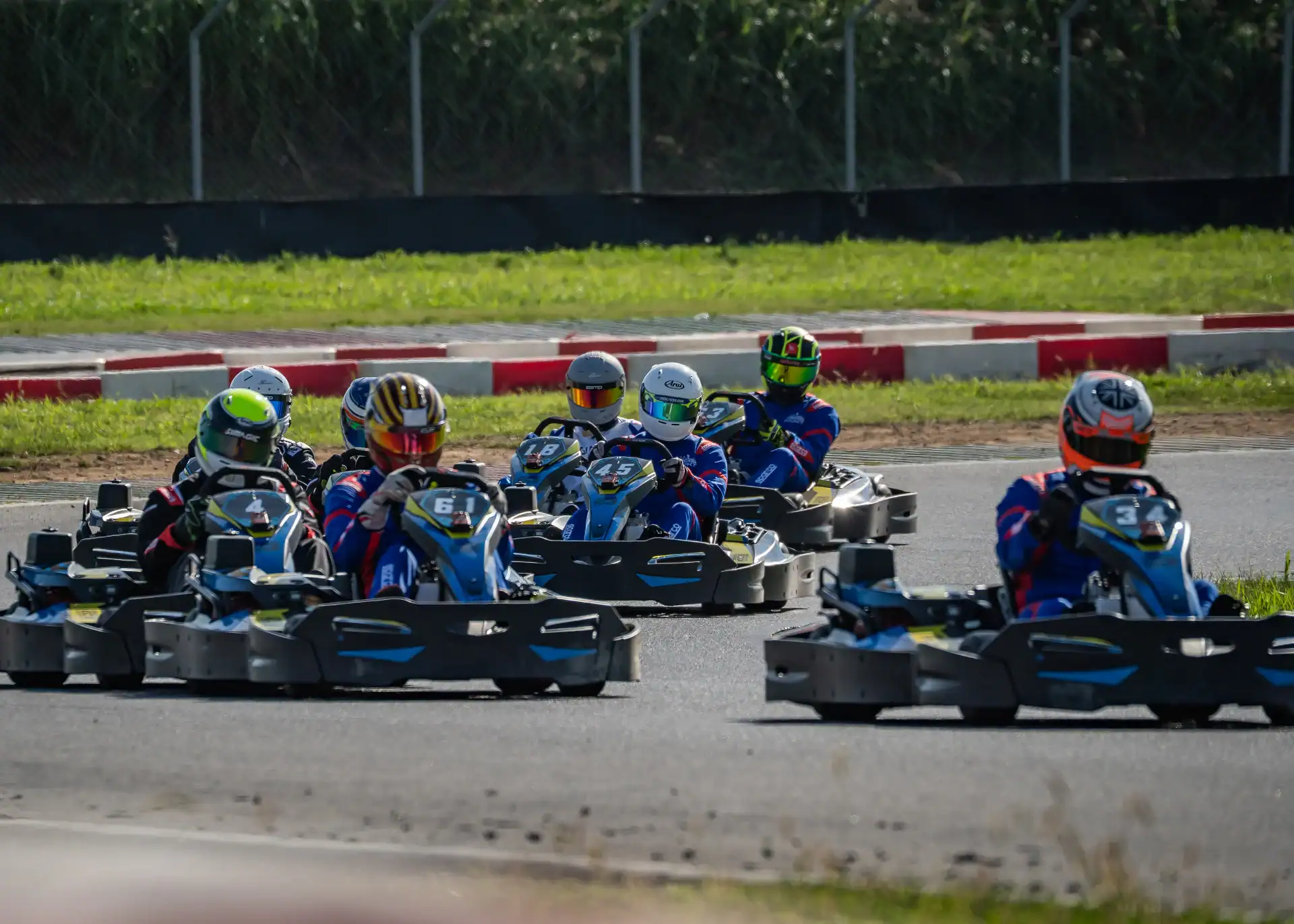 A group of go-kart racers in helmets and race suits drive closely together on an outdoor circuit.