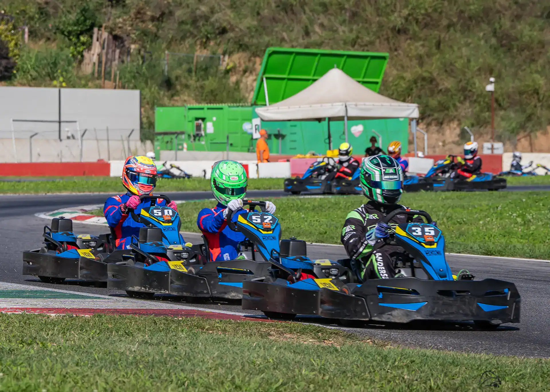 Three go-kart drivers in helmets and racing suits compete on an outdoor track, closely following each other round a bend.