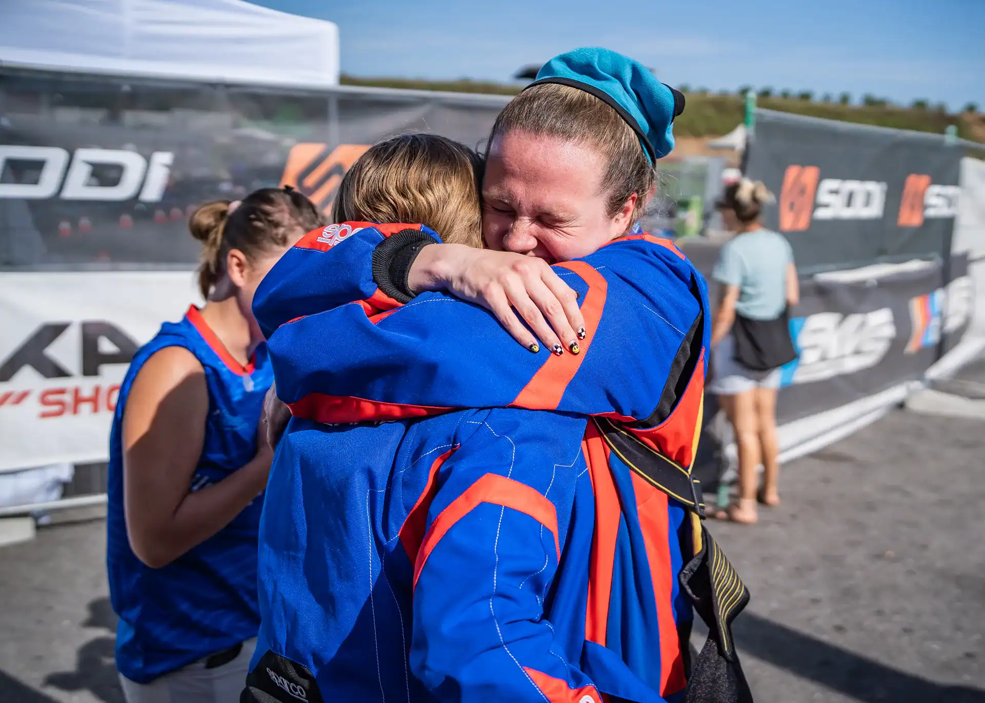 Two people wearing blue racing suits hug each other tightly at an outdoor event whilst others stand nearby.