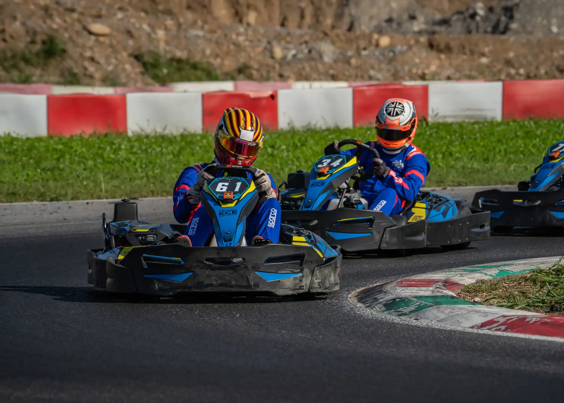 Two go-kart racers in blue suits and helmets compete on an outdoor track, closely rounding a bend bordered by red, white, and green kerbs.