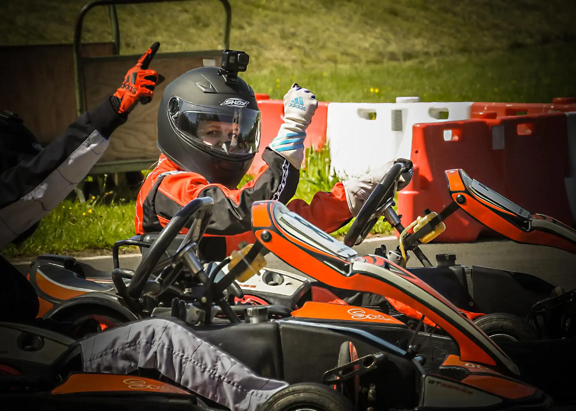 Person in a racing suit and helmet gives a thumbs-up whilst sitting in a go-kart on an outdoor track, with another racer and safety barriers visible nearby.