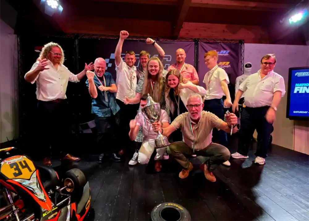 A group of people celebrates indoors around a trophy and a go-kart, with several raising their arms and smiling at the camera.