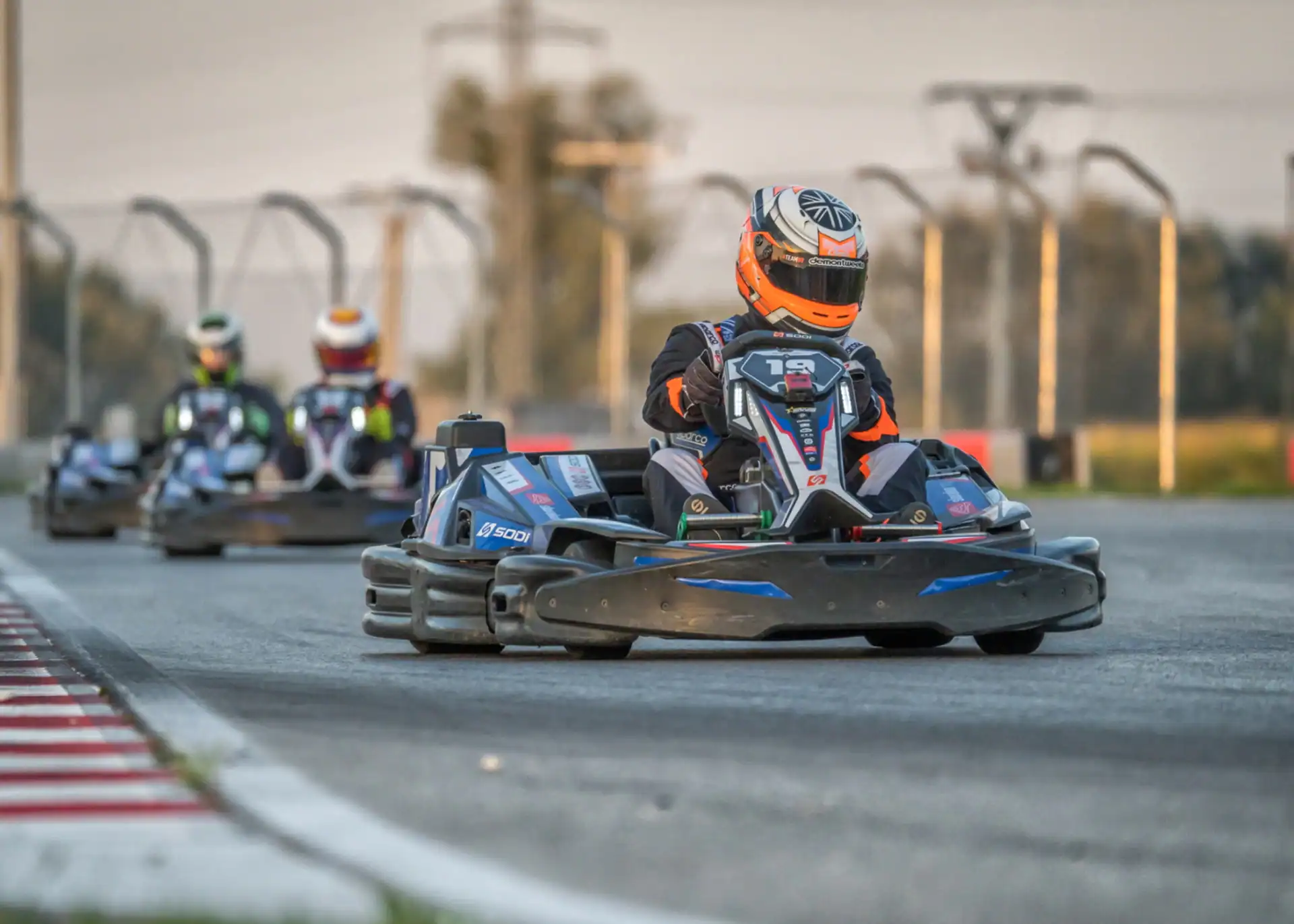 Three go-kart drivers race on a track, with the leading driver wearing an orange and black helmet and suit, closely followed by two competitors.