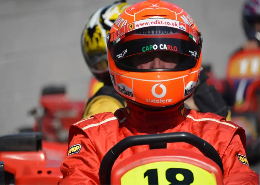 A racing car driver in a red suit and helmet sits in a vehicle marked with the number 18; other racers and karts are visible in the background.