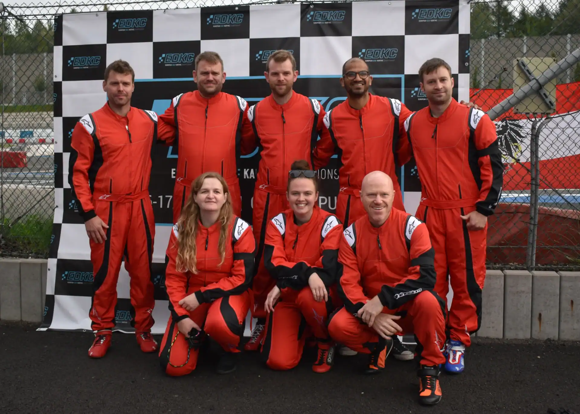 Eight people in matching red racing suits pose in front of a black and white chequered backdrop at a racetrack. Four are standing, four are crouching in front.