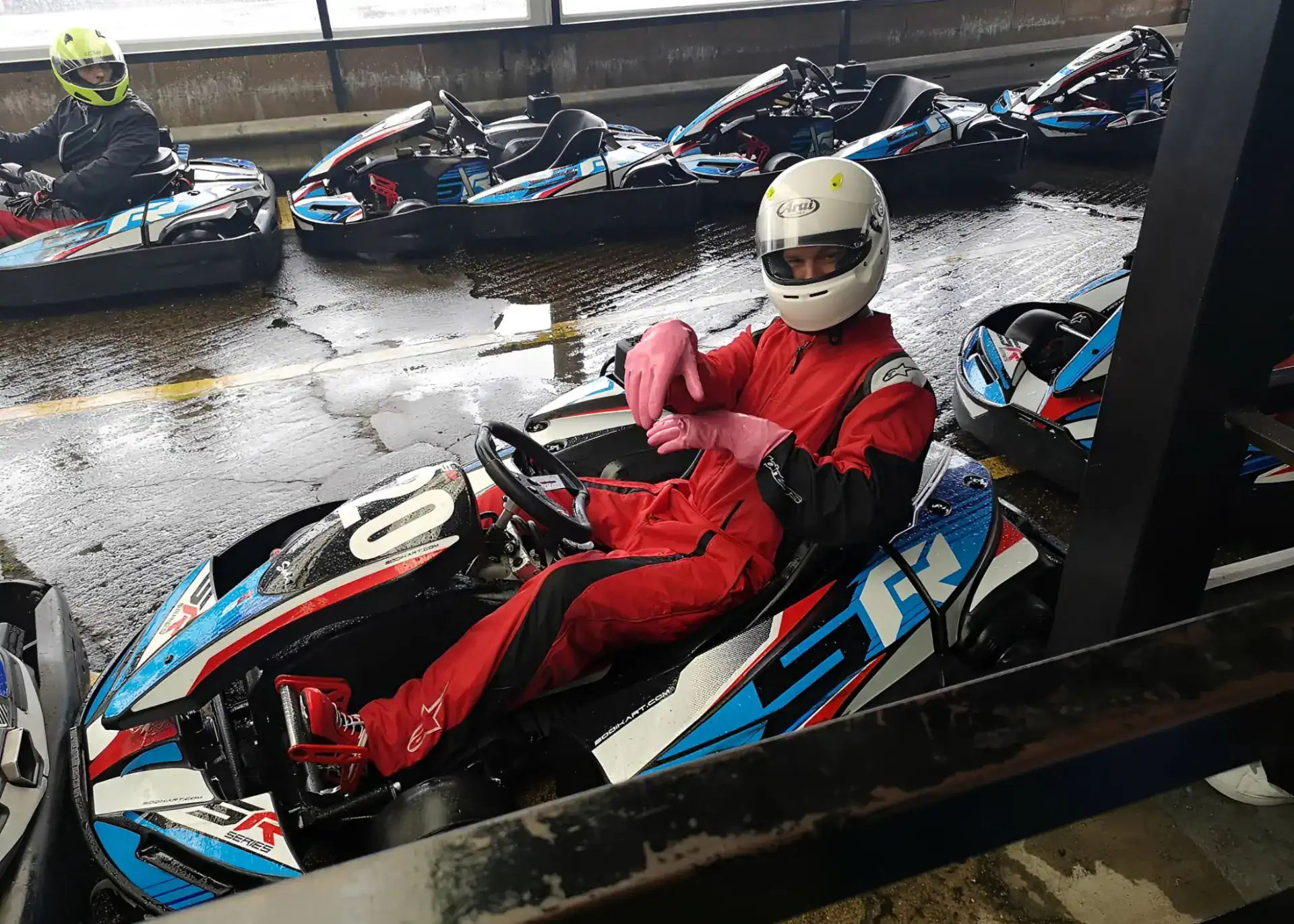 Person in a red racing suit and white helmet sits in a go-kart, wearing pink gloves, on a wet track with other go-karts and a driver in the background.