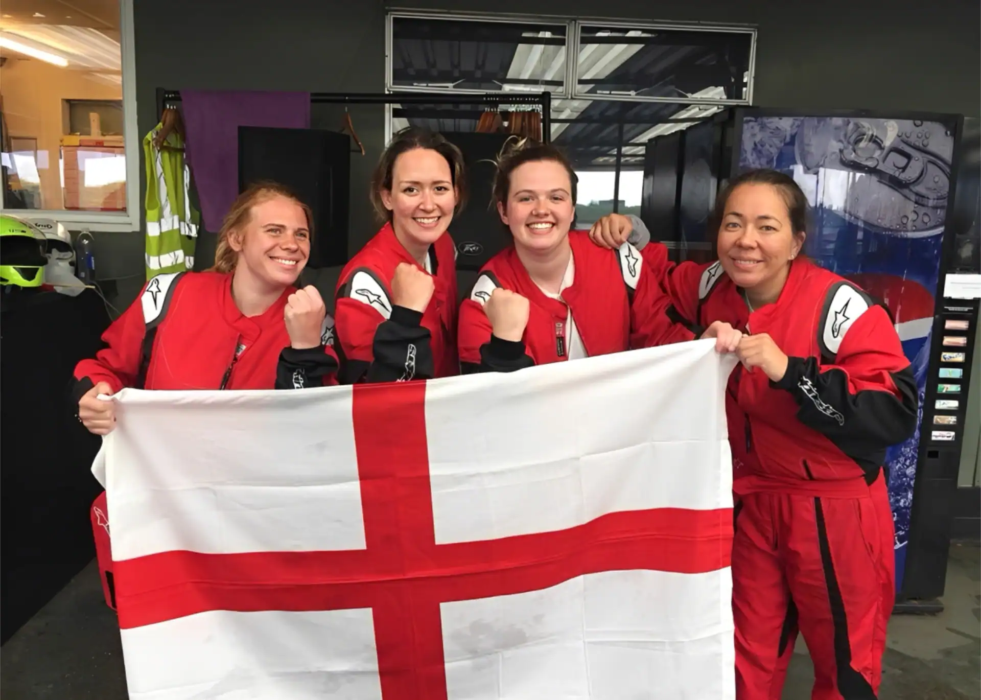 Four women in red racing suits hold a St George’s Cross flag and pose with raised fists, standing indoors near a vending machine and safety equipment.