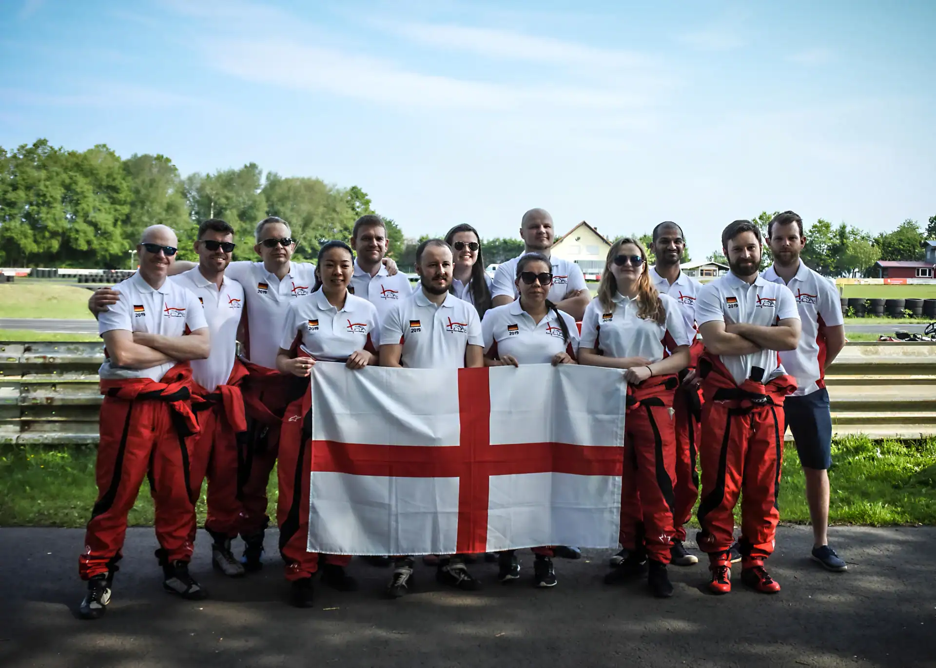 A group of people wearing white shirts and red trousers stand outdoors holding a large flag with a red cross on a white background.